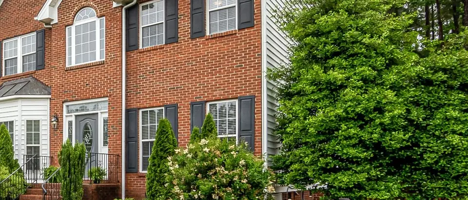 A two-storey red brick house with dark shutters and brick front steps behind a trimmed lawn, with tall pines to one side