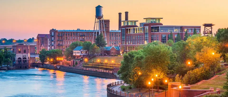 Red brick mill buildings and a steel water tower above the Chattahoochee River and the lamp-lit brick RiverWalk in Columbus, Georgia at dusk