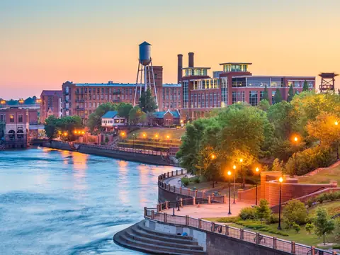Red brick mill buildings and a steel water tower above the Chattahoochee River and the lamp-lit brick RiverWalk in Columbus, Georgia at dusk