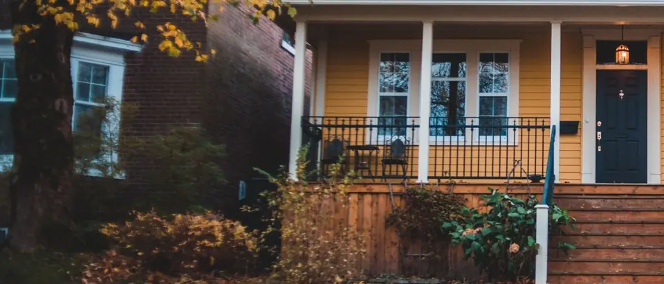 A yellow bungalow with a deep front porch and white posts above stone garden walls, with fallen leaves on the path and a brick house beside it