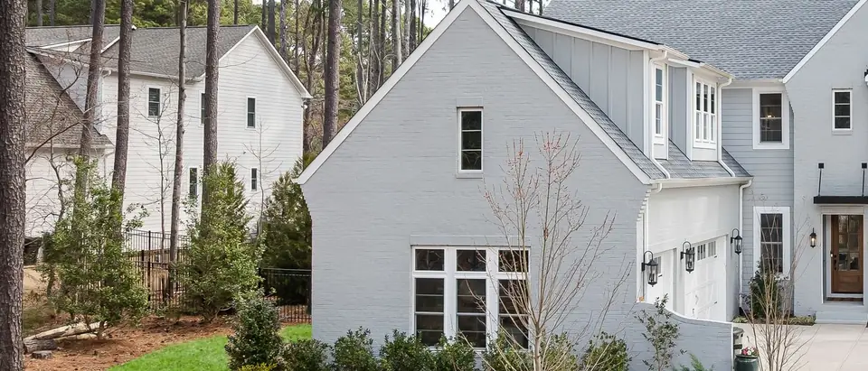 A pale grey two-storey house among tall pines, with a curved lawn, pine straw beds and a second house behind